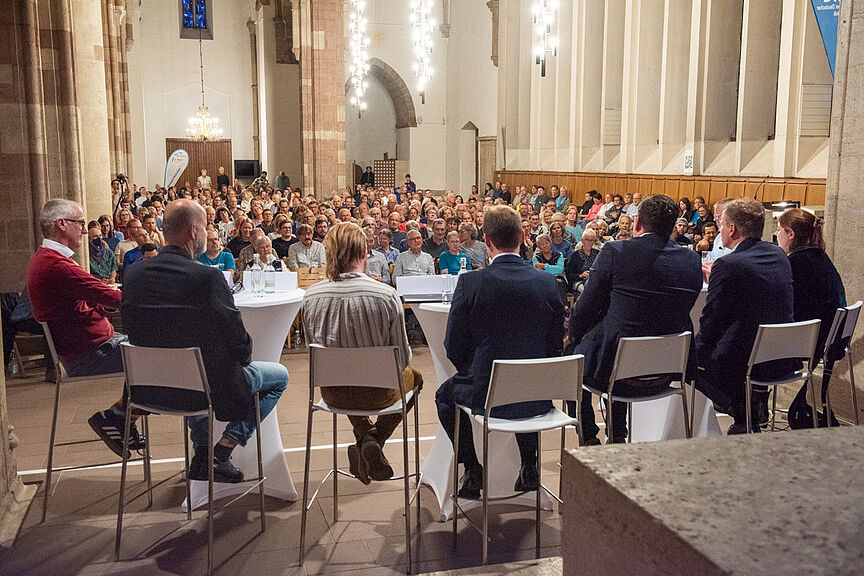 Podiumsdiskussion zur OB-Wahl Das Bild zeigt ein Podium mit sieben Personen und einen mit Menschen gefüllten Kirchenraum
