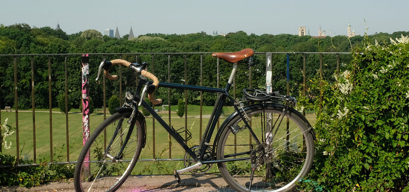 Das Bild zeigt im Vordergrund ein Fahrrad, im Hintergrund eine Wiese, Sportanlagen und Kirchtürme der Stadt