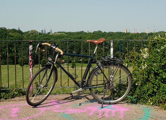 Das Bild zeigt im Vordergrund ein Fahrrad, im Hintergrund eine Wiese, Sportanlagen und Kirchtürme der Stadt