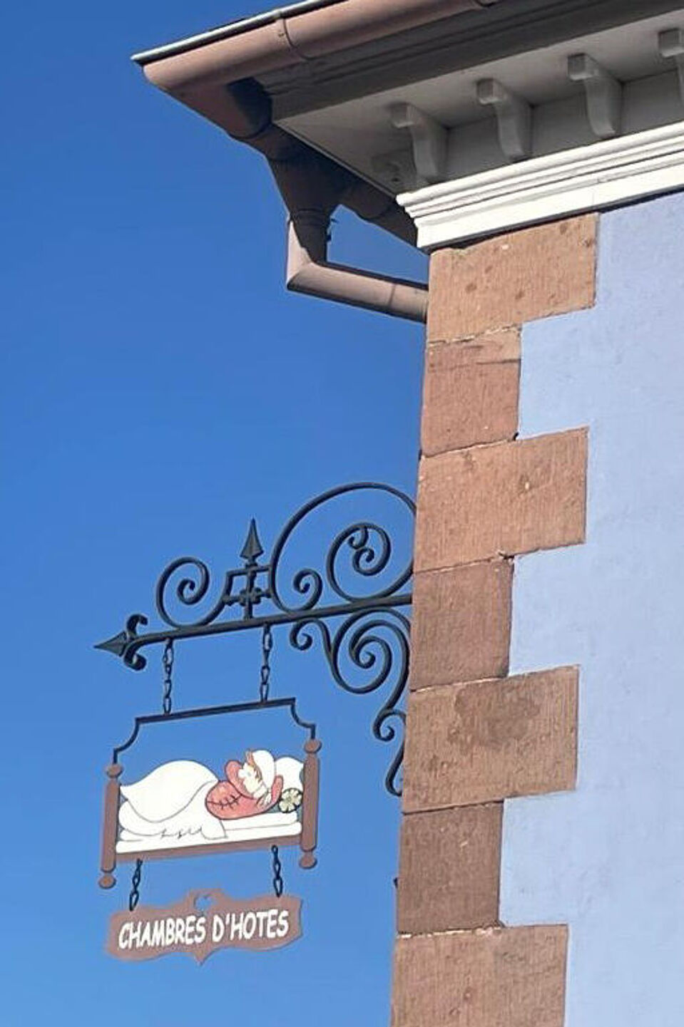 Chambres d'hotes im Elass Das Bild zeigt die Gebäudeecke eines Gasthauses im Elsass mit einem Schild; darauf ein Bett und die Beschriftung: Chambres d'hotes