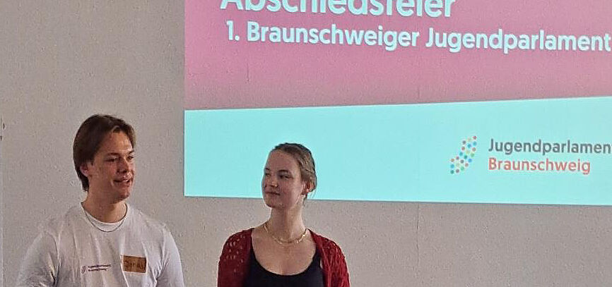 Das Bild zeigt zwei junge Menschen in einem Konferenzraum; auf der Projektionswand steht: Abschiedsfeier - 1. Braunschweiger Jugendparlament