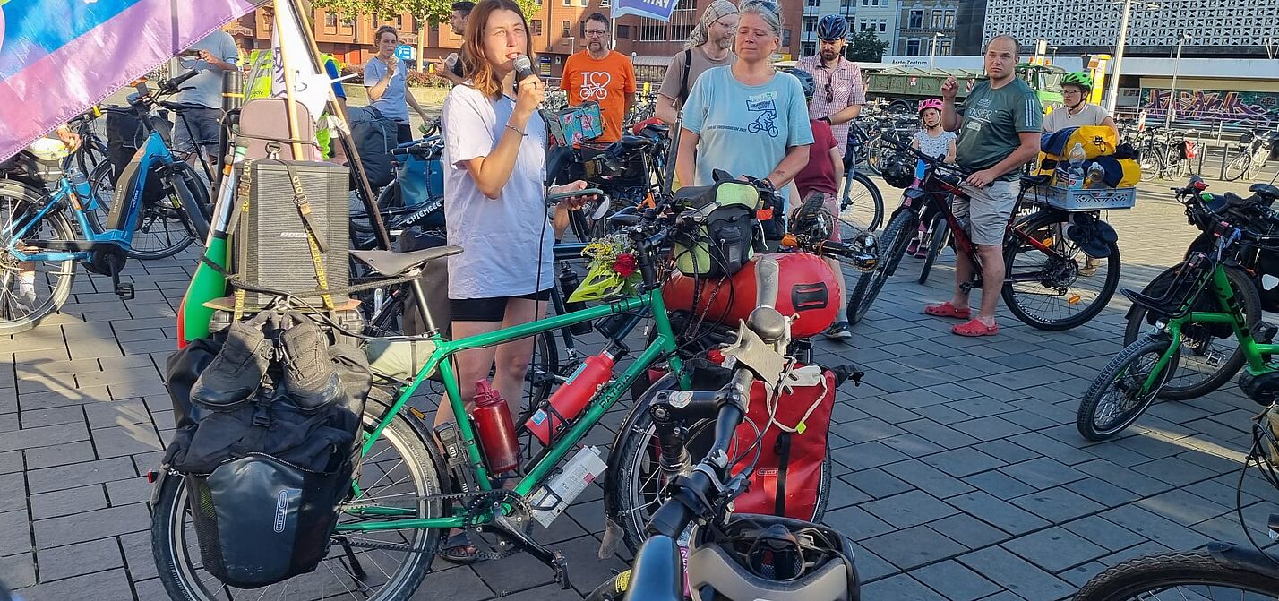 Das Bild zeigt eine Gruppe von Radlern, die sich auf einem Platz versammelt haben; eine junge Frau hat ein Mikrofon in der Hand
