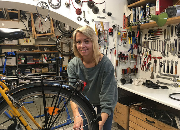 Das Bild zeigt eine junge Frau, die in einer Fahrradwerkstatt an einem Rad arbeitet