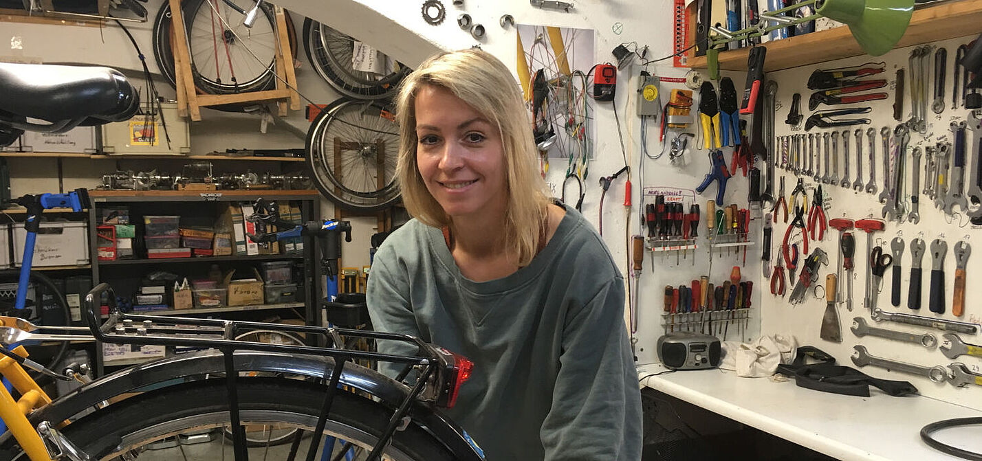Das Bild zeigt eine junge Frau, die in einer Fahrradwerkstatt an einem Rad arbeitet